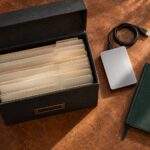An archive box of folders beside an external backup drive and green notebook on a copper-coloured desk.