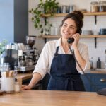 A café proprietor in a navy apron answers a corded telephone behind the counter.
